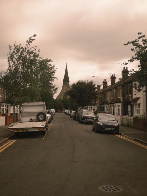 A residential street scene during daytime with parked cars lining both sides of the road, which is paved and features double yellow lines along the curb. In the foreground, a white van with an open re