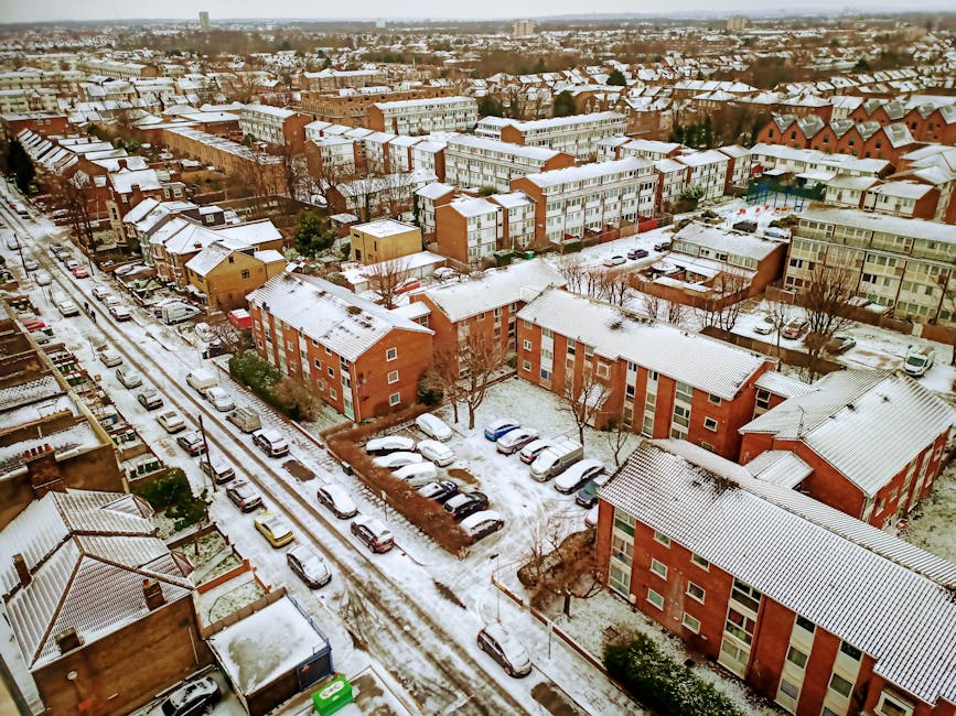 An aerial view of a residential area during winter showing multiple multi-storey apartment buildings and townhouses with snow-covered roofs. A narrow street runs through the scene, lined with parked c